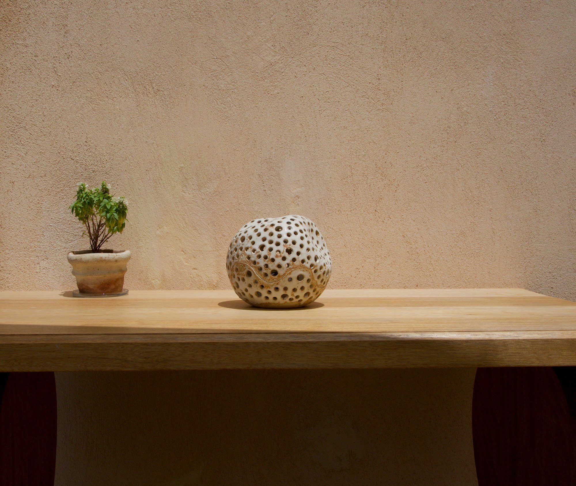 An oak table against a plaster wall, with a small bonsai and a pierced ceramic sphere