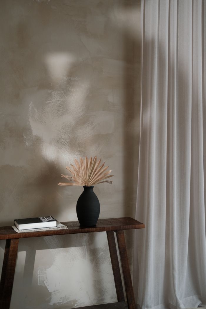 A small wooden console against a pale plaster wall, a black vase of dried grasses and a book
