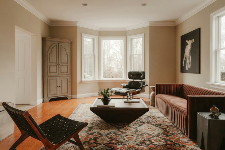A living room of pieces chosen one by one: a rust velvet sofa, a leather lounge chair in the bay window, a woven sling chair, an antique cupboard and a Persian rug