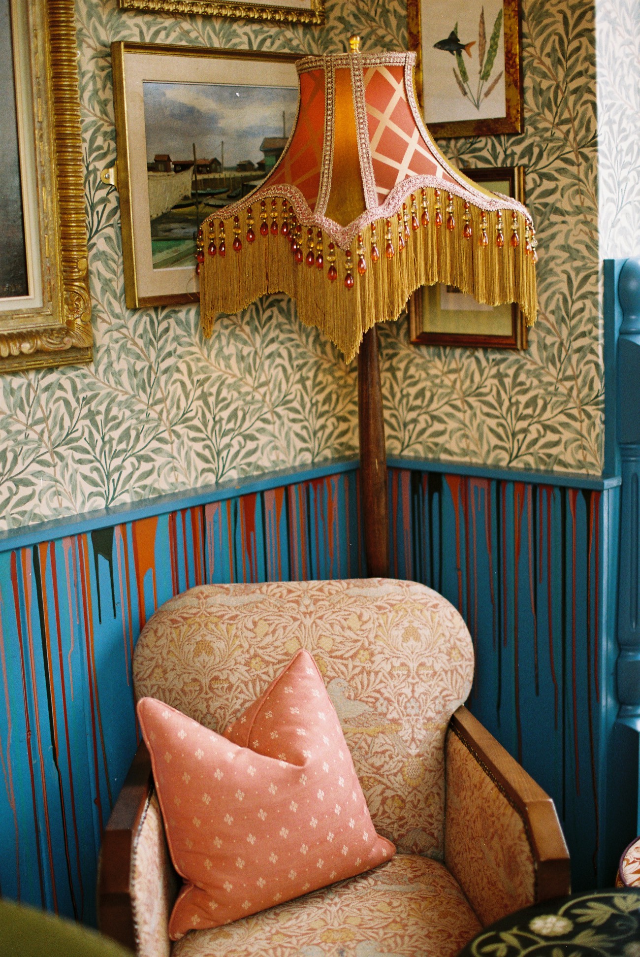 A richly layered corner: botanical wallpaper above striped panelling, a fringed lampshade, a landscape oil painting and a patterned armchair