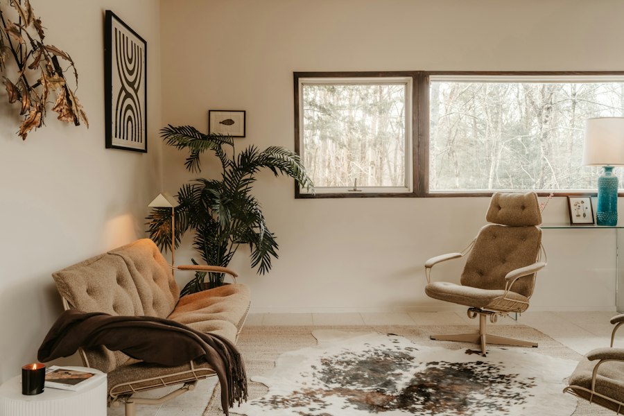 A collected sitting room: a tufted tan sofa and a matching swivel lounge chair on a cowhide, a palm, framed prints and a long window onto trees