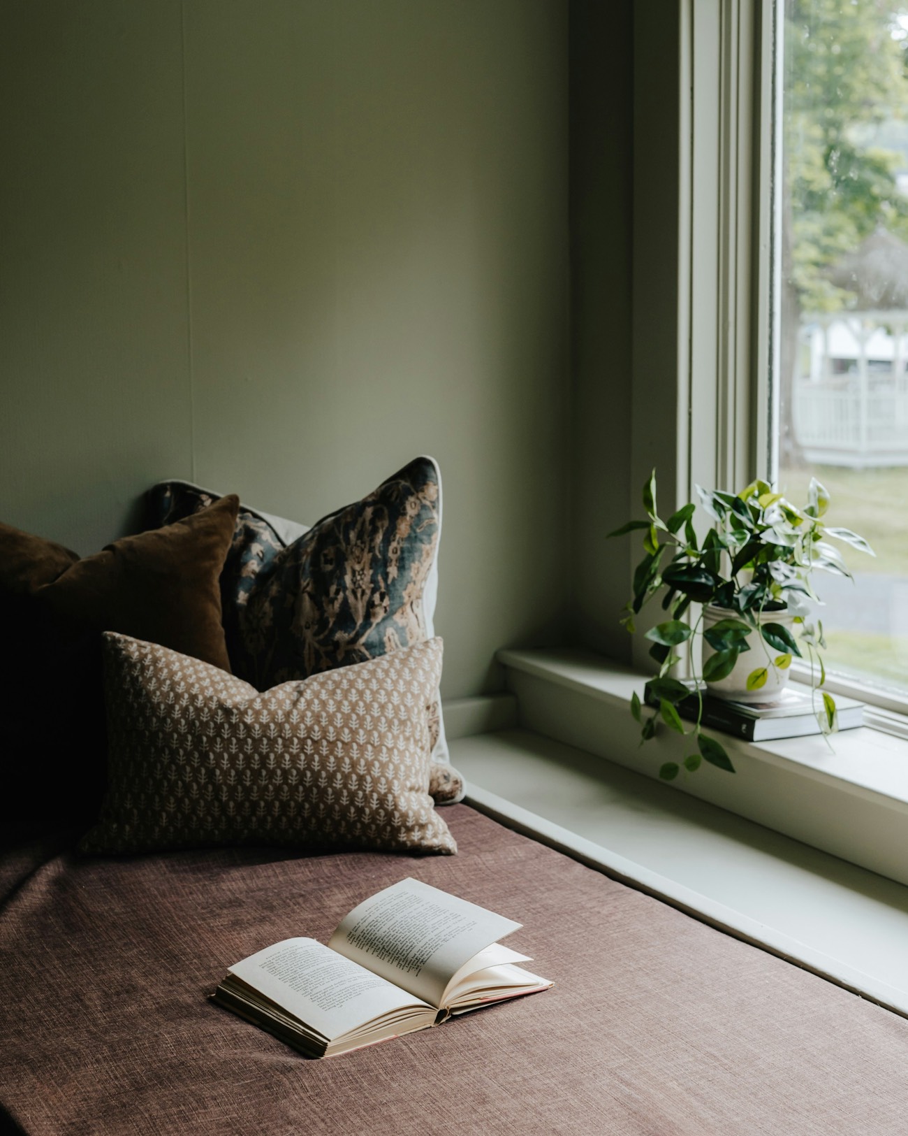 A window seat with linen and patterned cushions, an open book and a trailing plant in soft window light