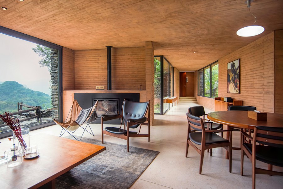 A rammed-earth house with a board-formed ceiling: a sculptural leather armchair and a sling chair by a steel fireplace, a teak dining table, glass onto green hills