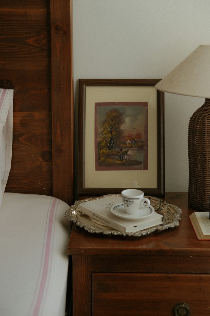 A bedside table with a small framed landscape, a teacup on a silver tray and a rattan lamp