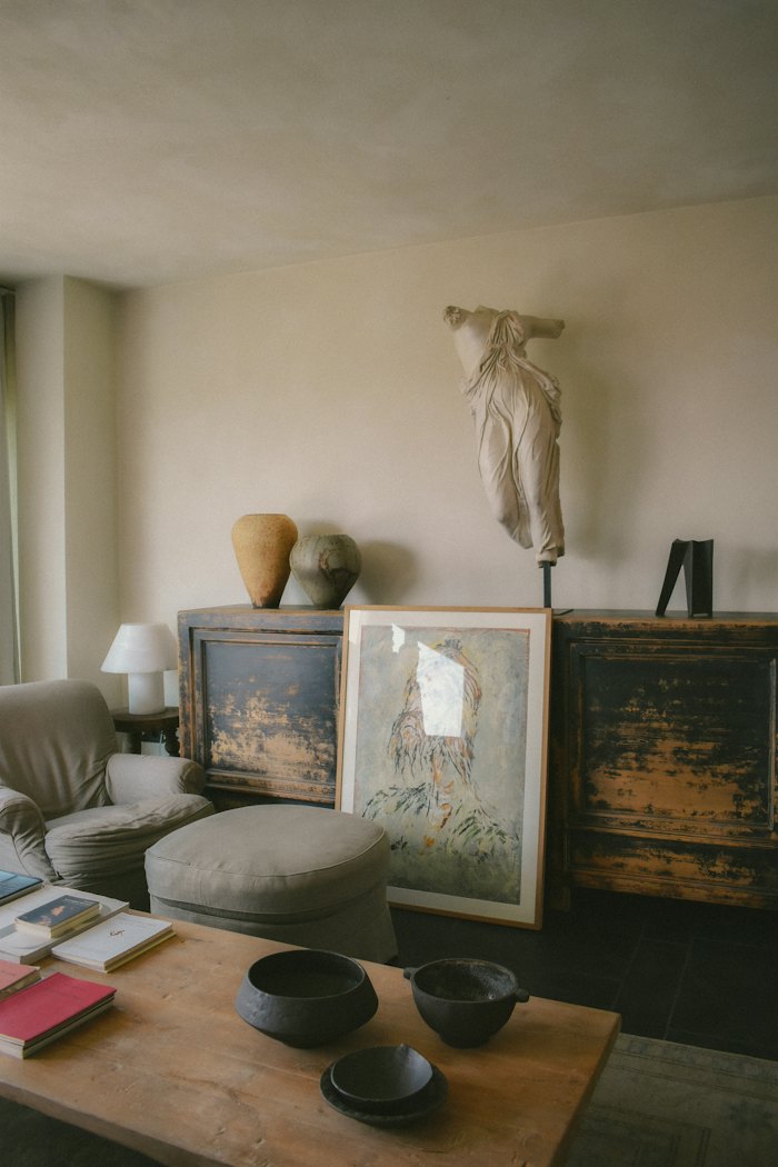 A lived-in sitting room: a painted cabinet with a classical torso and stoneware vases, a framed print, bowls and books on a low table