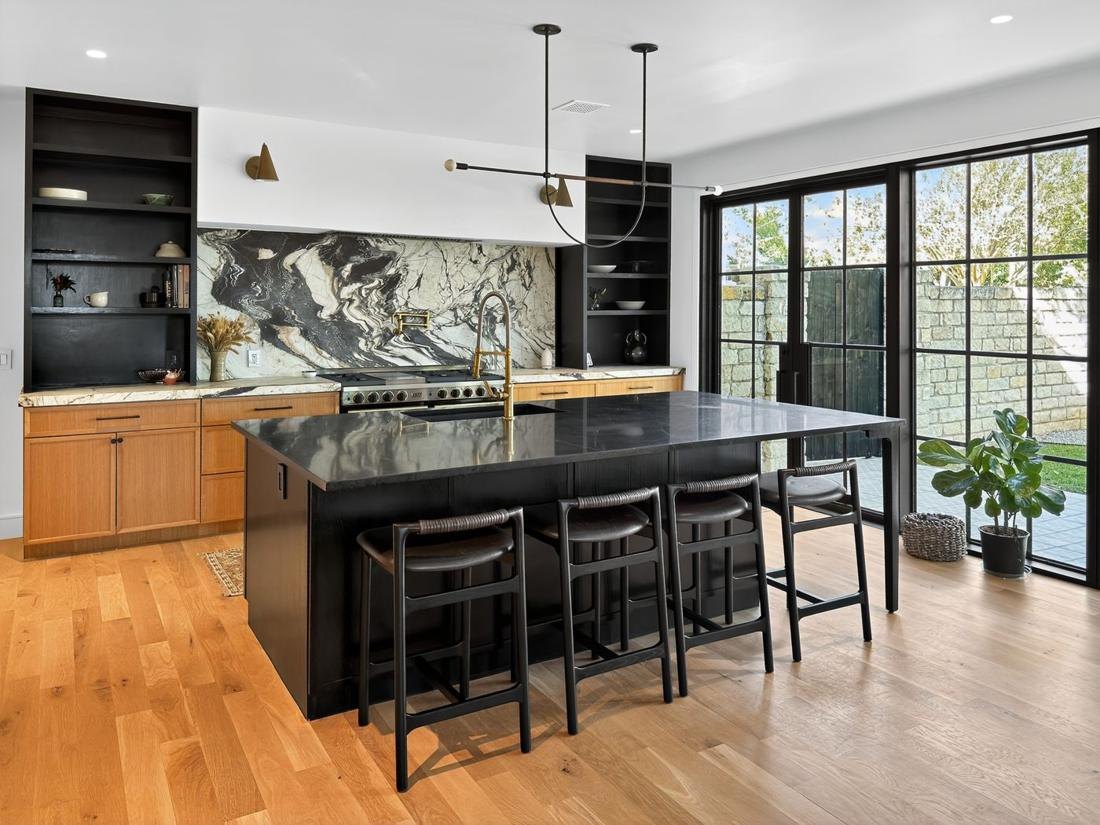 A kitchen with a black island, a marble backsplash to the ceiling and a wall of wood cabinetry