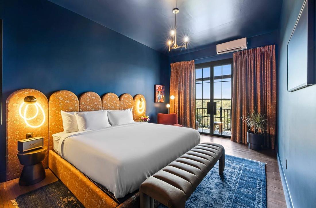 A hotel room with a deep blue ceiling, a scalloped velvet headboard and doors onto a balcony