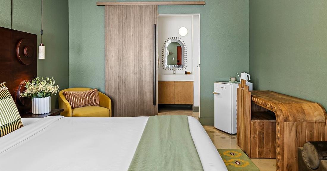 A hotel room with sage walls, a carved wood desk and a sliding door to a bath with an arched mirror